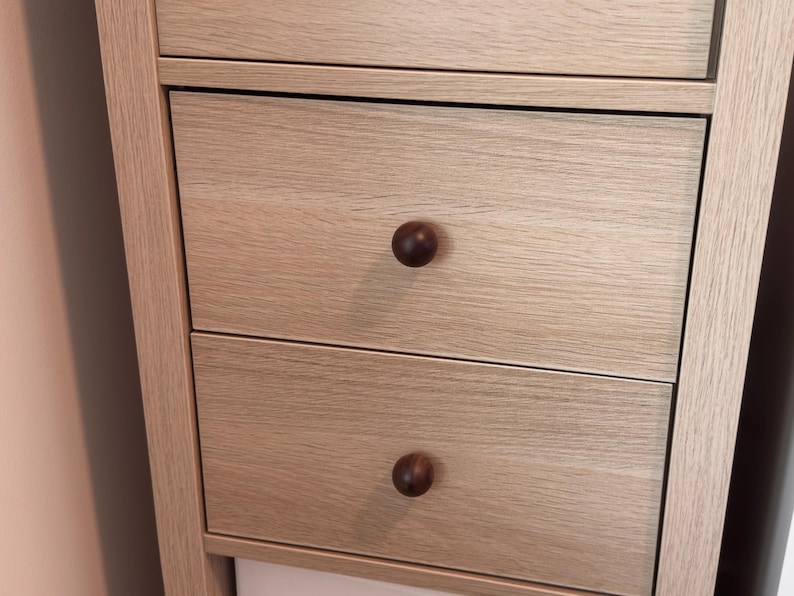 May include: Close-up of a light-coloured wooden cabinet with three drawers. Each drawer has a dark brown, round knob. The cabinet has a simple, modern design with visible wood grain.