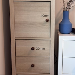 May include: A light-coloured wooden cabinet with a door and two drawers. The door has a dark brown knob, and the drawers have dark brown knobs. The door knob is 40mm in diameter, and the drawer knobs are 30mm in diameter. A blue vase and a white cabinet are in the background.