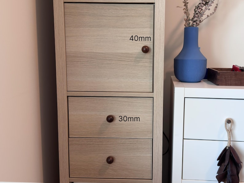 May include: A light-coloured wooden cabinet with a door and two drawers. The door has a dark brown knob, and the drawers have dark brown knobs. The door knob is 40mm in diameter, and the drawer knobs are 30mm in diameter. A blue vase and a white cabinet are in the background.