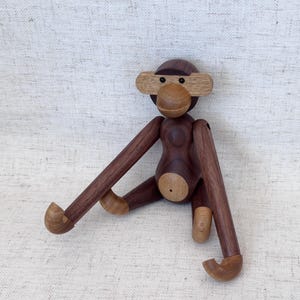 May include: A wooden monkey figurine crafted from dark and light-toned wood. The monkey features a rounded head with a horizontal light wood band across the eyes, a circular snout, a smooth torso, and long, articulated limbs with curved ends.