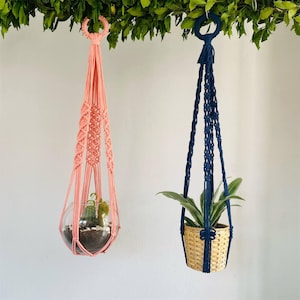 Puede incluir: Dos colgadores de plantas de macramé. Uno es de color rosa coral, con un terrario de cristal con cactus. El otro es azul marino, con una cesta tejida con una planta verde. Ambos están suspendidos de una rama verde y frondosa.