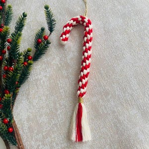 Könnte beinhalten: Handgefertigter Weihnachtsbaumschmuck in Form einer Zuckerstange. Die Zuckerstange besteht aus rot-weiß geflochtenem Kordel, mit einem goldfarbenen Akzent an der Basis und einer Quaste. Ein Zweig mit roten Beeren ist im Hintergrund.