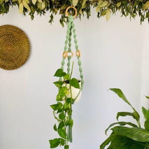 Könnte beinhalten: Ein hellgrüner Makramee-Pflanzenaufhänger mit Holzringen, der einen weißen Keramiktopf mit einer grünen Blattpflanze hält. Der Pflanzenaufhänger ist an einem Holzring aufgehängt.