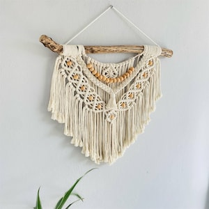 Op de afbeelding: Crèmekleurige macramé wandhanger met een geometrisch ontwerp, houten kralen en franjes. De macramé is opgehangen aan een natuurlijke drijfhouttak. Een decoratief stuk voor een bohemian interieur.