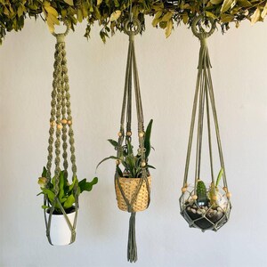 Puede incluir: Tres colgadores de plantas de macramé en verde oliva, suspendidos de una rama con hojas verdes. Uno sostiene una maceta blanca, otro una cesta tejida y el tercero un terrario de vidrio. Cada uno presenta cuentas de madera y un diseño único.