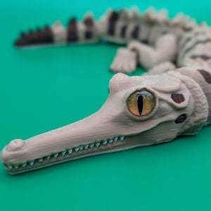May include: An articulated crocodile figurine with a light beige body, dark brown spots, and a long snout. The creature features a prominent, detailed eye with a yellow iris and a vertical black pupil, along with visible teeth and segmented body parts.