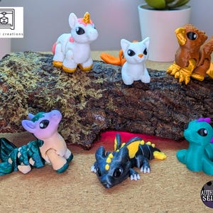 May include: A collection of colorful 3D-printed fantasy creatures, including a unicorn, cat, and various dragons, displayed on a textured wooden surface. The figures are in shades of white, blue, yellow, and orange.
