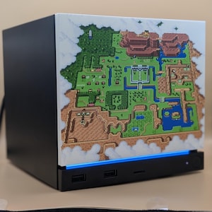 Könnte beinhalten: Ein schwarzes Computergehäuse mit einer weißen Frontplatte, die eine farbenfrohe 16-Bit-Pixel-Art-Kartenillustration zeigt. Die Basis enthält einen horizontalen blauen LED-Lichtstreifen, zwei USB-Anschlüsse und einen runden Netzschalter.