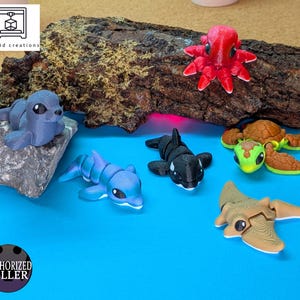 Puede incluir: Una colección de criaturas marinas impresas en 3D, que incluye un pulpo rojo, un delfín azul, una orca en blanco y negro, una tortuga verde y marrón, una manta raya beige y una foca gris. Los artículos se exhiben sobre una superficie azul con un trozo de madera.
