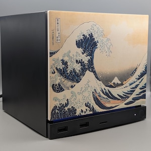 Pode incluir: Uma caixa de computador preta com um painel frontal impresso com a obra de arte da Grande Onda de Kanagawa. O design apresenta grandes ondas azuis, um pequeno pico de montanha e caligrafia japonesa tradicional num fundo de cor creme.
