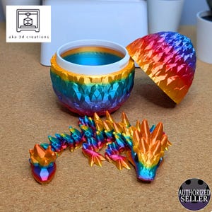 Könnte beinhalten: Ein mehrfarbiger, 3D-gedruckter Drache und Behälter. Der Drache ist beweglich und hat einen Regenbogenfarbverlauf. Der Behälter ist kugelförmig mit abnehmbarem Deckel, ebenfalls in einem Regenbogenverlauf. Der Text "aka 3d creations" ist sichtbar.