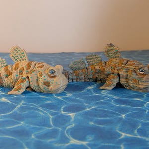 May include: Two articulated fish figurines with segmented bodies. The left fish is tan with brown spots and teal accents. The right fish is brown with darker brown stripes. Both feature large, prominent eyes and detailed fin structures.