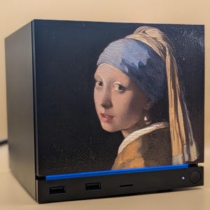 Könnte beinhalten: Ein schwarzes, rechteckiges elektronisches Gerät mit einer Reproduktion des Gemäldes Das Mädchen mit dem Perlenohrgehänge auf der Vorderseite. Das Gerät verfügt über einen blauen Lichtstreifen, drei USB-Anschlüsse und einen runden Einschaltknopf.
