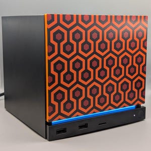 May include: A black computer case featuring a front panel with a repeating orange, red, and dark brown hexagonal geometric pattern. The base includes a horizontal blue light strip, two USB ports, a small slot, and a circular power button on the right.