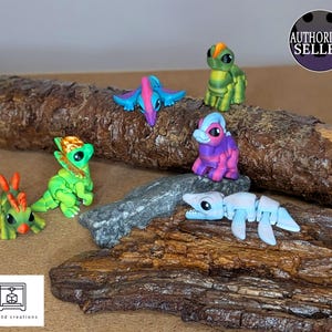 May include: A collection of colorful 3D-printed fantasy creatures, including dragons and other mythical beings. The figures are in shades of green, blue, purple, and orange, and are arranged on a piece of driftwood and a rock. The image includes the text "AUTHORIZED SELLER".