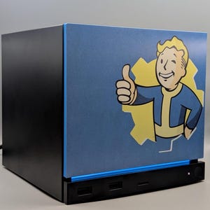 Könnte beinhalten: Ein schwarzes Computergehäuse mit einer blauen Frontplatte, die eine Grafik des Fallout Vault Boy Charakters mit erhobenem Daumen zeigt. Die Figur ist in Gelb und Blau vor einem gelben, zahnradförmigen Hintergrund auf der blauen Fläche dargestellt.