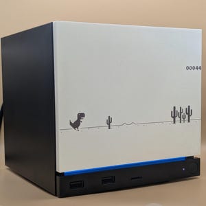 May include: A black and off-white computer case featuring a pixelated dinosaur and cactus graphic on the side panel. The front panel includes a blue light strip, three USB ports, and the number 00044 printed in black on the upper right corner.