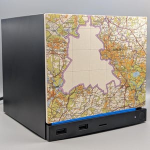 May include: A black computer case featuring a side panel with a detailed topographic map of Berlin and Potsdam. The map displays green and orange terrain colors with a purple outline highlighting the city area. A blue accent strip sits below.