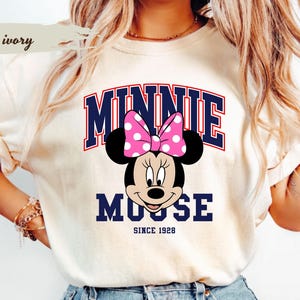May include: Ivory t-shirt with a Minnie Mouse graphic, including a pink polka-dot bow. The word "MINNIE" is arched in navy blue, with "MOOSE" below, and "SINCE 1928" at the bottom. The shirt is a light cream colour.