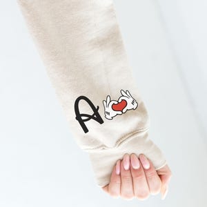 May include: Beige sweatshirt sleeve with the letter "A" in black and a cartoon hand heart graphic with a red heart. The sleeve is on a person's arm.