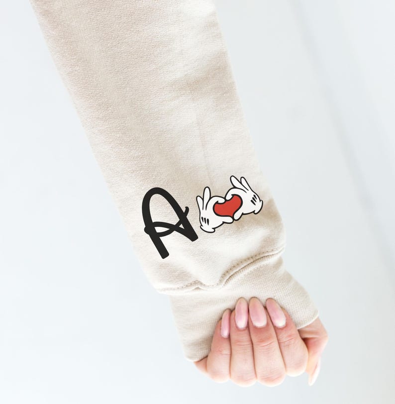 May include: Beige sweatshirt sleeve with the letter "A" in black and a cartoon hand heart graphic with a red heart. The sleeve is on a person's arm.