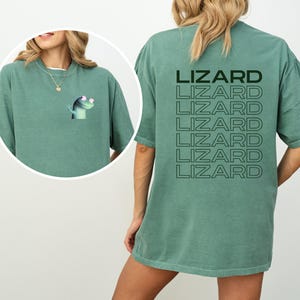 May include: A sage green t-shirt featuring a cartoon lizard graphic on the front and the word "LIZARD" repeated vertically on the back. The lizard is light green with pink accents. The shirt has a relaxed fit.