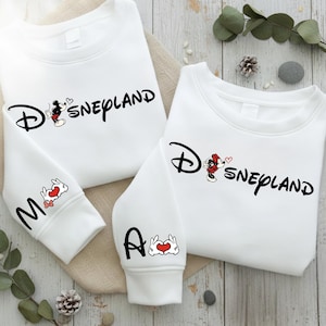 May include: White sweatshirts with the word "Disneyland" in black script, featuring Mickey Mouse and a heart. One sleeve has the letter "M" and a rabbit with a heart, the other has the letter "A" and a hand-heart symbol.