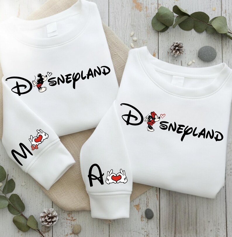 May include: White sweatshirts with the word "Disneyland" in black script, featuring Mickey Mouse and a heart. One sleeve has the letter "M" and a rabbit with a heart, the other has the letter "A" and a hand-heart symbol.