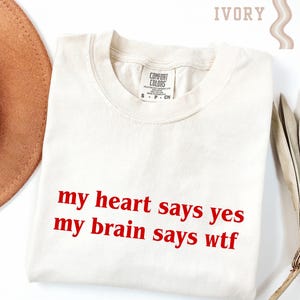 May include: Ivory-colored t-shirt with the text "my heart says yes my brain says wtf" in red. The shirt is folded and has a Comfort Colors tag. A brown hat and decorative leaves are partially visible.