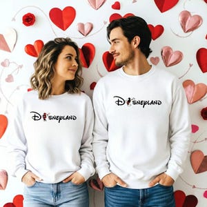 May include: Two white sweatshirts with the word "Disneyland" in black script. The "D" in Disneyland is stylized with the Disney logo. The sweatshirts are worn by a couple in front of a wall decorated with red and pink hearts.