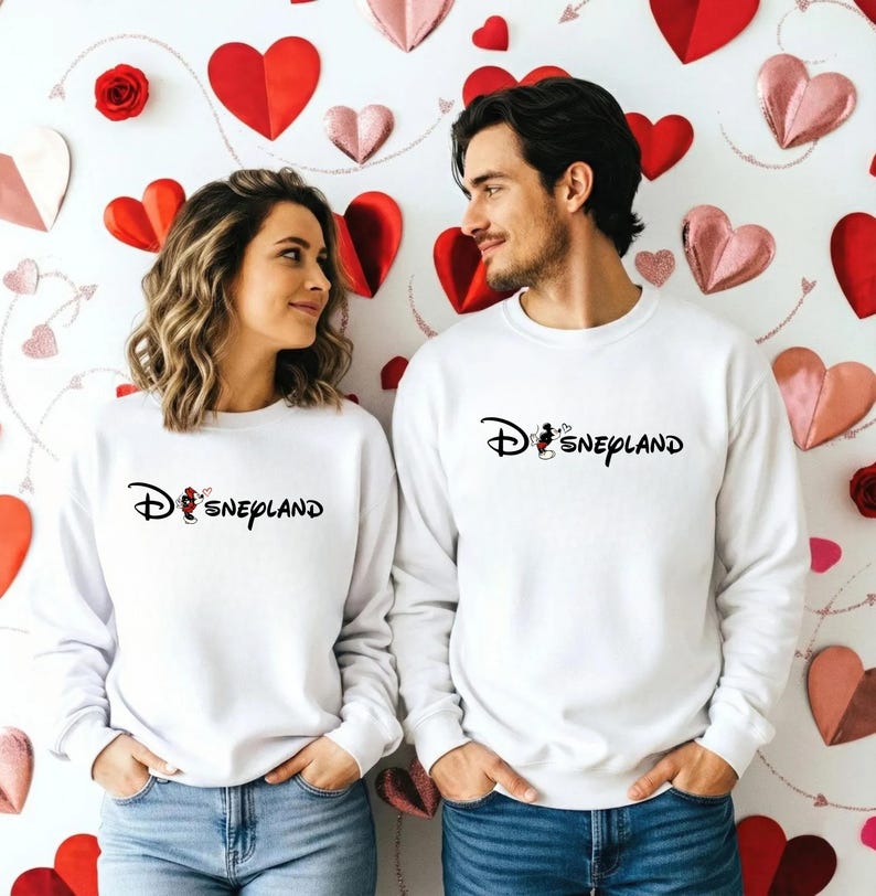 May include: Two white sweatshirts with the word "Disneyland" in black script. The "D" in Disneyland is stylized with the Disney logo. The sweatshirts are worn by a couple in front of a wall decorated with red and pink hearts.
