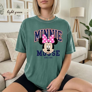 May include: A light green t-shirt with a Minnie Mouse graphic. The design includes a pink polka dot bow, the word "MINNIE" in red and navy blue, and "MOUSE" below. The text "SINCE 1928" is at the bottom of the graphic.