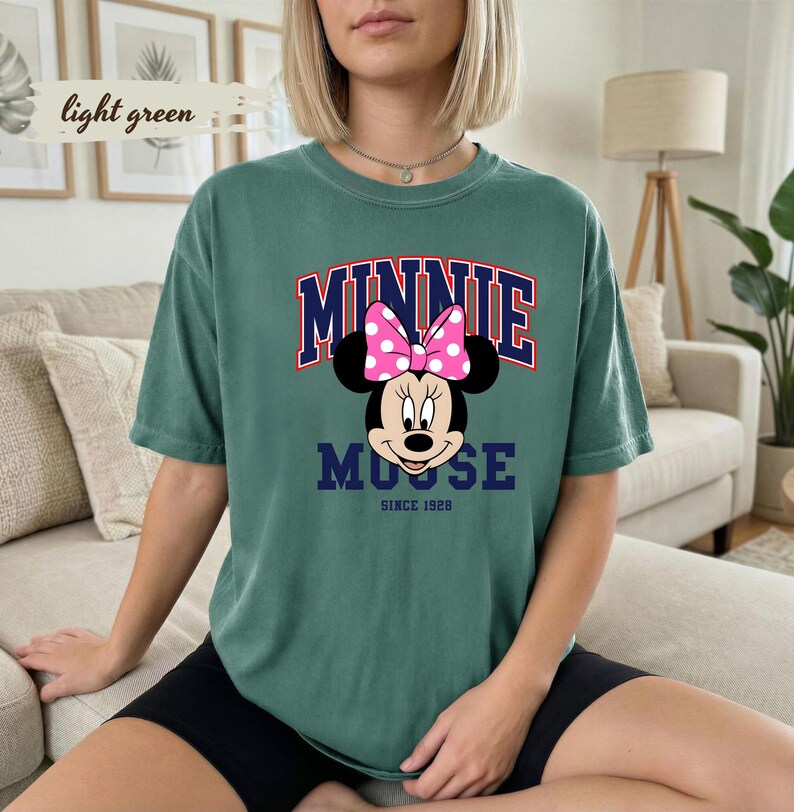 May include: A light green t-shirt with a Minnie Mouse graphic. The design includes a pink polka dot bow, the word "MINNIE" in red and navy blue, and "MOUSE" below. The text "SINCE 1928" is at the bottom of the graphic.