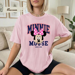 May include: A pale pink t-shirt with a Minnie Mouse graphic. The design includes a pink polka dot bow and the text "MINNIE MOUSE" above the character, with "SINCE 1928" below. The shirt has a relaxed fit.
