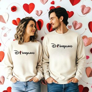 May include: Two people wearing matching cream-colored sweatshirts with the word "Disneyland" in black script. The sweatshirts feature a Mickey Mouse graphic. The background is decorated with red and pink paper hearts.