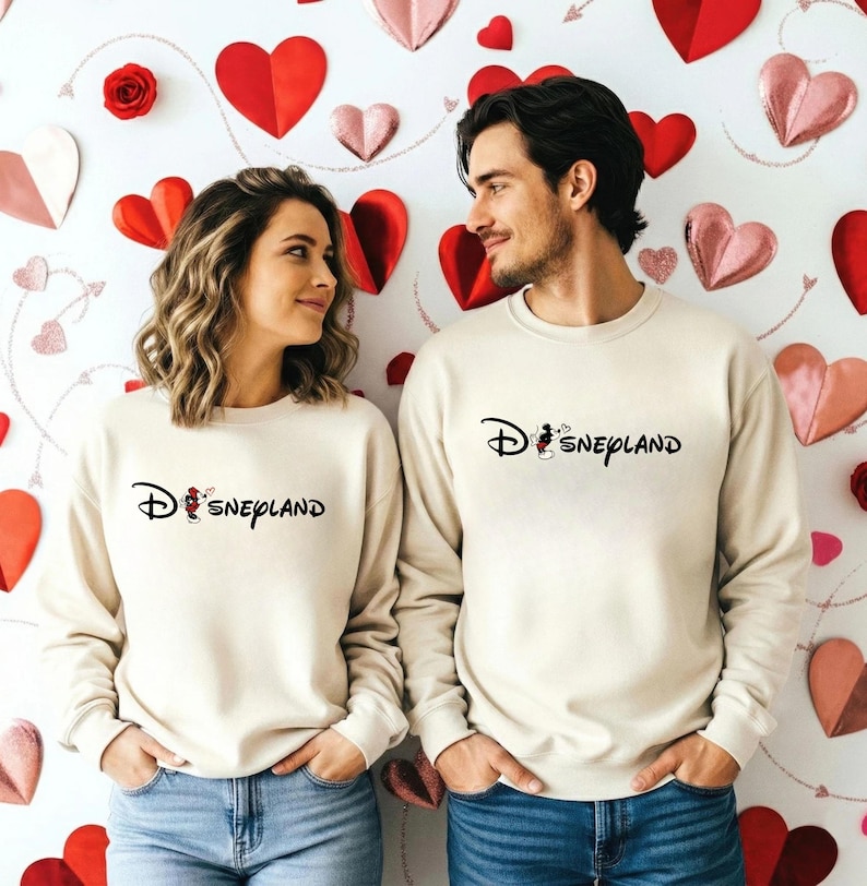 May include: Two people wearing matching cream-colored sweatshirts with the word "Disneyland" in black script. The sweatshirts feature a Mickey Mouse graphic. The background is decorated with red and pink paper hearts.