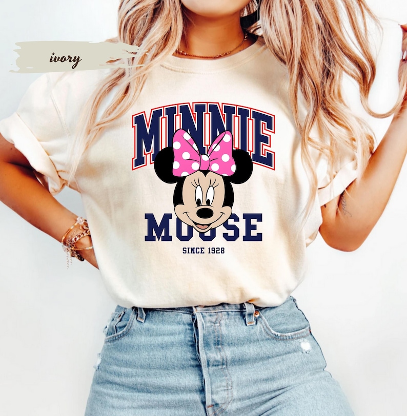 May include: Ivory t-shirt with a Minnie Mouse graphic, including a pink polka-dot bow. The word "MINNIE" is arched in navy blue, with "MOOSE" below, and "SINCE 1928" at the bottom. The shirt is a light cream colour.