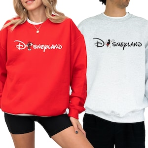 May include: Red and gray sweatshirts with the word "Disneyland" in a script font. The "D" in Disneyland is stylized with a Mickey Mouse graphic. The red sweatshirt is worn with black shorts. The gray sweatshirt is worn with black pants.
