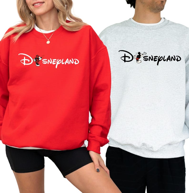 May include: Red and gray sweatshirts with the word "Disneyland" in a script font. The "D" in Disneyland is stylized with a Mickey Mouse graphic. The red sweatshirt is worn with black shorts. The gray sweatshirt is worn with black pants.