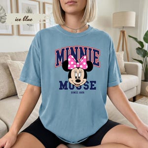May include: An ice blue t-shirt with a Minnie Mouse graphic. The design includes the word "MINNIE" in red and navy blue, above Minnie's face, and "MOUSE" below. A pink polka dot bow is featured. The text "SINCE 1928" is also included.