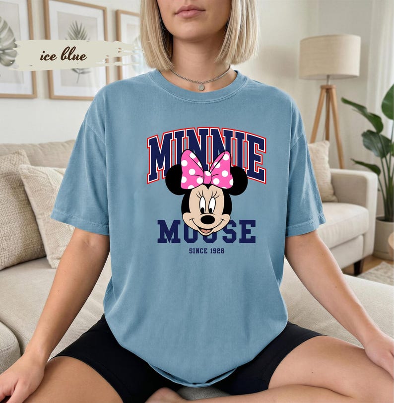 May include: An ice blue t-shirt with a Minnie Mouse graphic. The design includes the word "MINNIE" in red and navy blue, above Minnie's face, and "MOUSE" below. A pink polka dot bow is featured. The text "SINCE 1928" is also included.