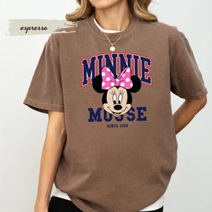 May include: Brown t-shirt with a Minnie Mouse graphic. The design features the name "MINNIE" in red and blue, a pink polka-dot bow, and the text "MOOSE SINCE 1928". The shirt has a relaxed fit and short sleeves.