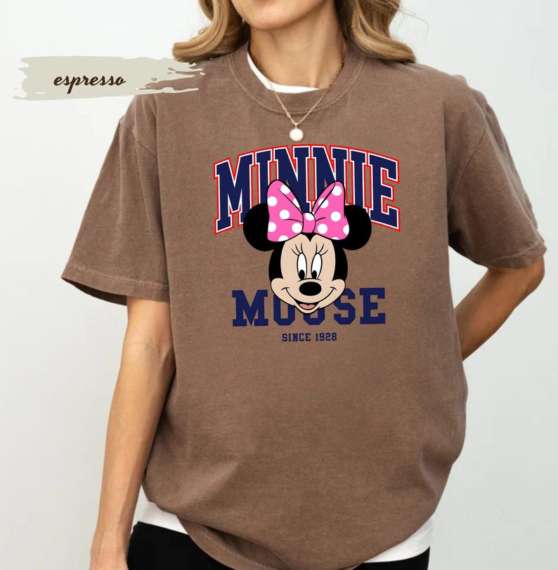 May include: Brown t-shirt with a Minnie Mouse graphic. The design features the name "MINNIE" in red and blue, a pink polka-dot bow, and the text "MOOSE SINCE 1928". The shirt has a relaxed fit and short sleeves.
