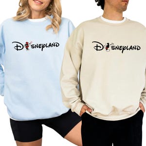 May include: Two sweatshirts, one light blue and one beige, each with the word "Disneyland" in a script font. The "D" is stylized with a Mickey Mouse graphic. The light blue sweatshirt is paired with black shorts.