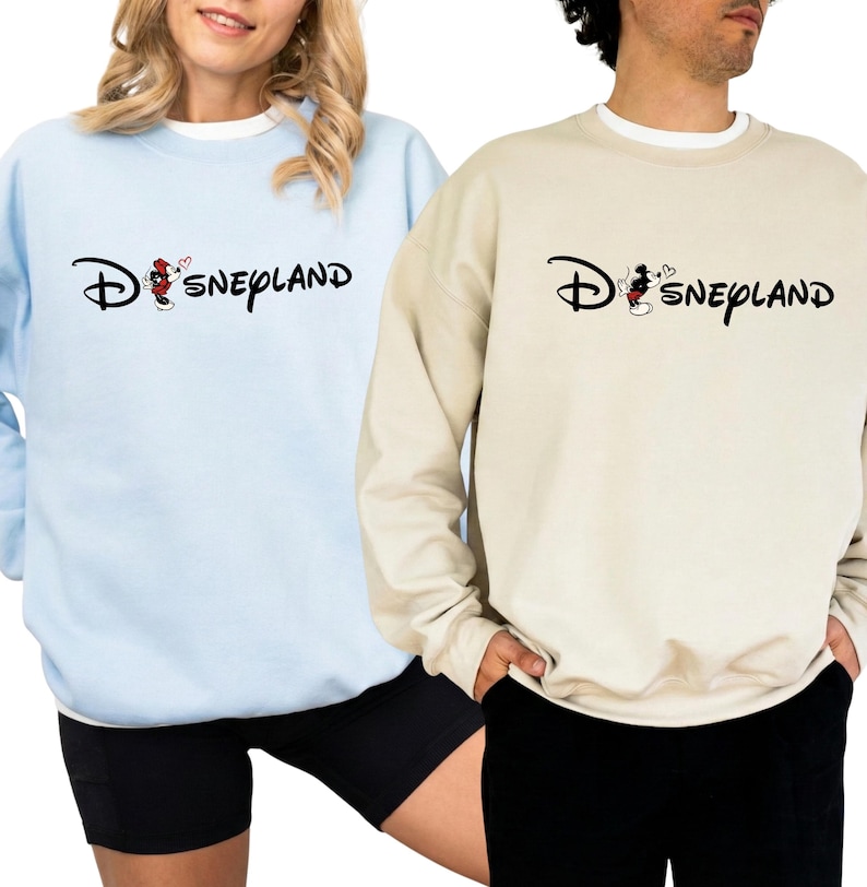 May include: Two sweatshirts, one light blue and one beige, each with the word "Disneyland" in a script font. The "D" is stylized with a Mickey Mouse graphic. The light blue sweatshirt is paired with black shorts.