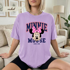 May include: A lilac t-shirt with a Minnie Mouse graphic, including a pink polka-dot bow. The text "MINNIE MOUSE" is arched across the top in navy and red, with "SINCE 1928" below. The shirt has a relaxed fit.