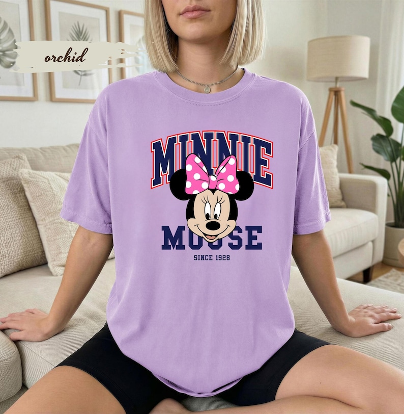May include: A lilac t-shirt with a Minnie Mouse graphic, including a pink polka-dot bow. The text "MINNIE MOUSE" is arched across the top in navy and red, with "SINCE 1928" below. The shirt has a relaxed fit.