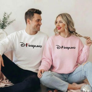 May include: A white and a pink sweatshirt with the word "Disneyland" printed in black. The "D" in Disneyland is stylized with a Mickey Mouse graphic. The white sweatshirt has a Mickey Mouse graphic with a red heart.