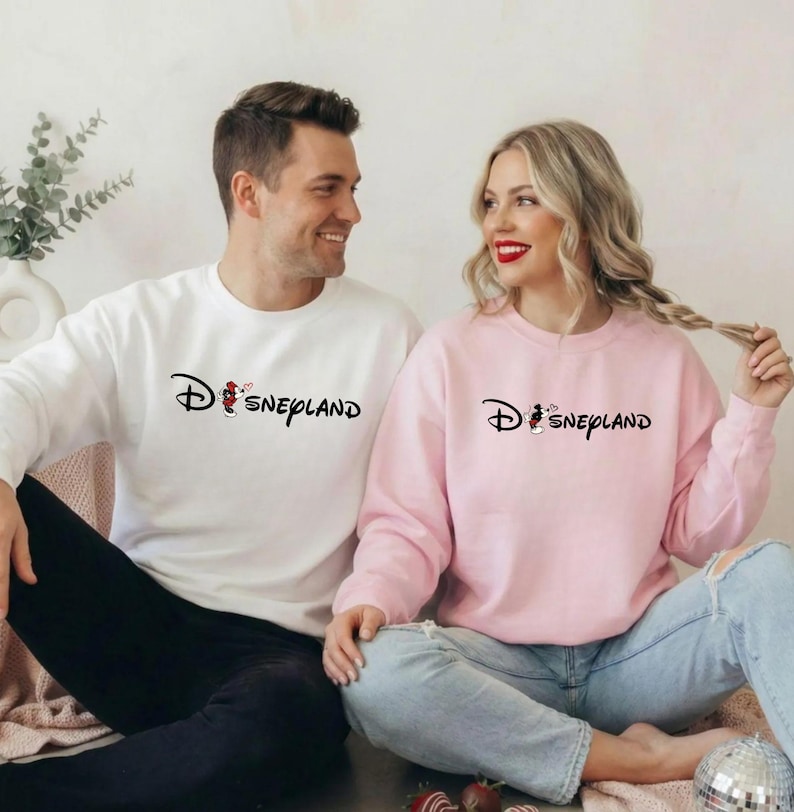 May include: A white and a pink sweatshirt with the word "Disneyland" printed in black. The "D" in Disneyland is stylized with a Mickey Mouse graphic. The white sweatshirt has a Mickey Mouse graphic with a red heart.