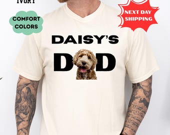 May include: Ivory-colored t-shirt with the text "DAISY'S DAD" in black block letters. The letter "D" features a photo of a brown and white dog. The shirt also has the text "COMFORT COLORS" and "NEXT DAY SHIPPING".